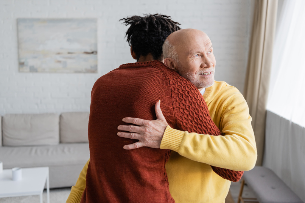 Cheerful senior man hugging african american grandson in living room  - Foto, afbeelding