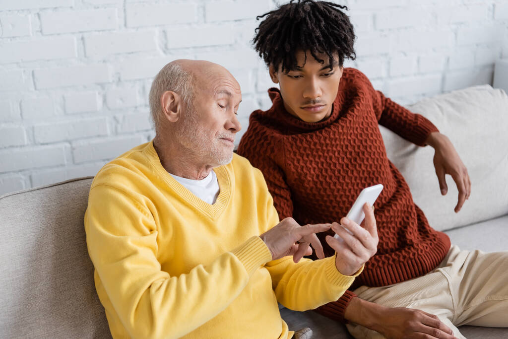 Пожилой человек указывает на смартфон рядом с африканским внуком на диване дома  - Фото, изображение