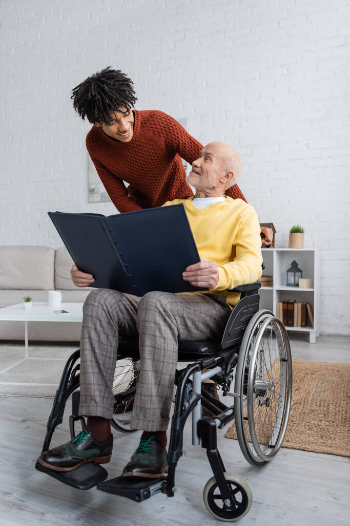 Positive pensioner holding photo album while sitting in wheelchair near african american grandson at home  - Foto, imagen