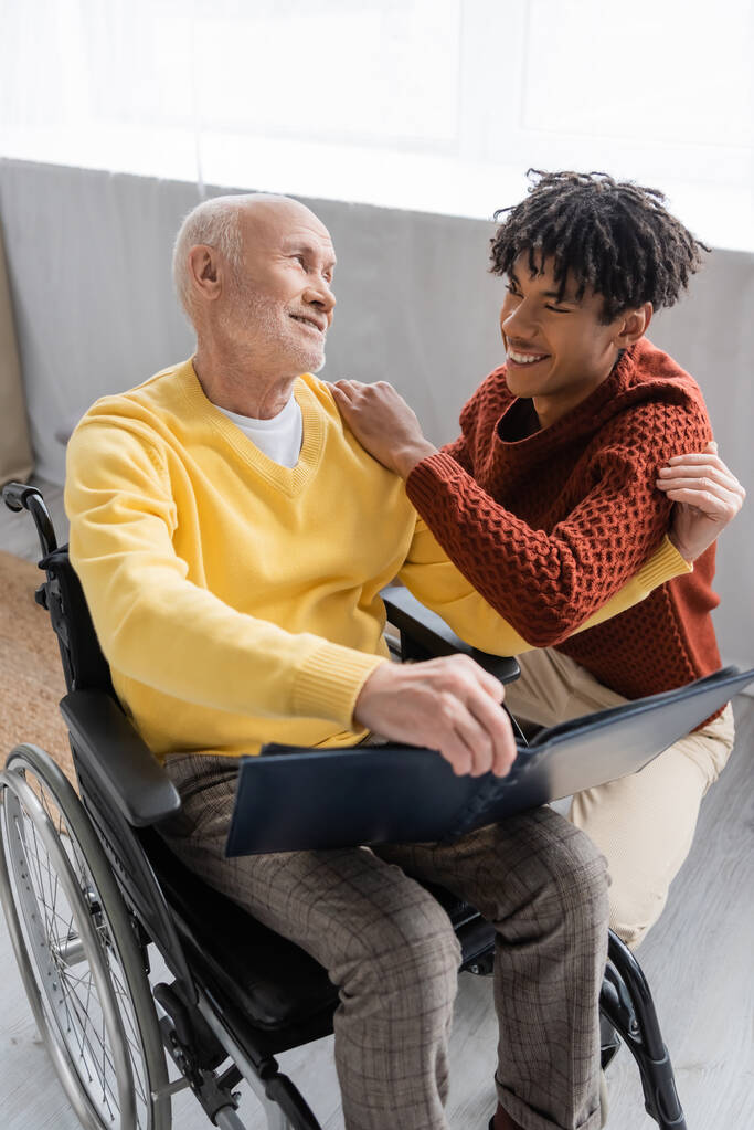 Позитивний афроамериканський онук обіймає дідуся з фотоальбомом у інвалідному візку вдома
  - Фото, зображення