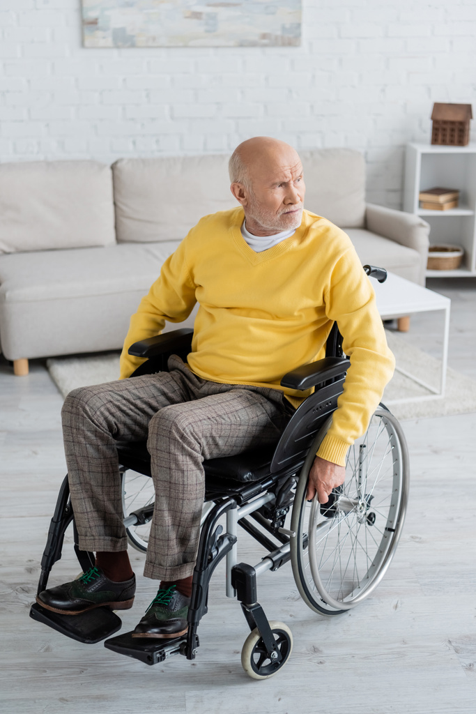Ηλικιωμένος άνδρας κοιτάζει αλλού, ενώ κάθεται σε αναπηρική καρέκλα στο σπίτι - Φωτογραφία, εικόνα
