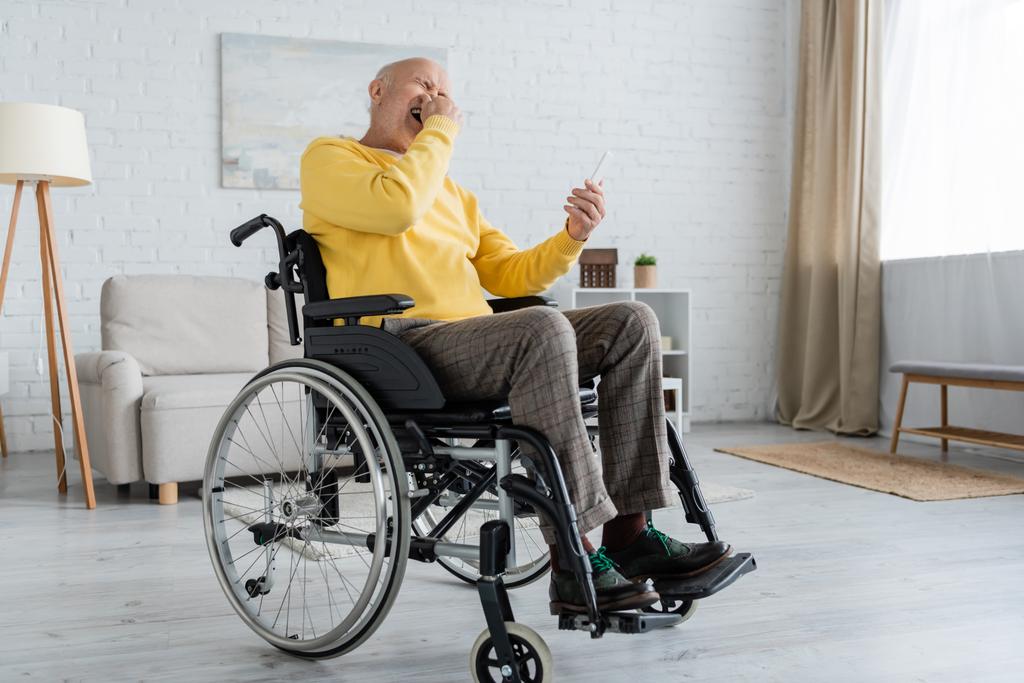 Laughing senior man holding smartphone while sitting in wheelchair at home  - Photo, Image