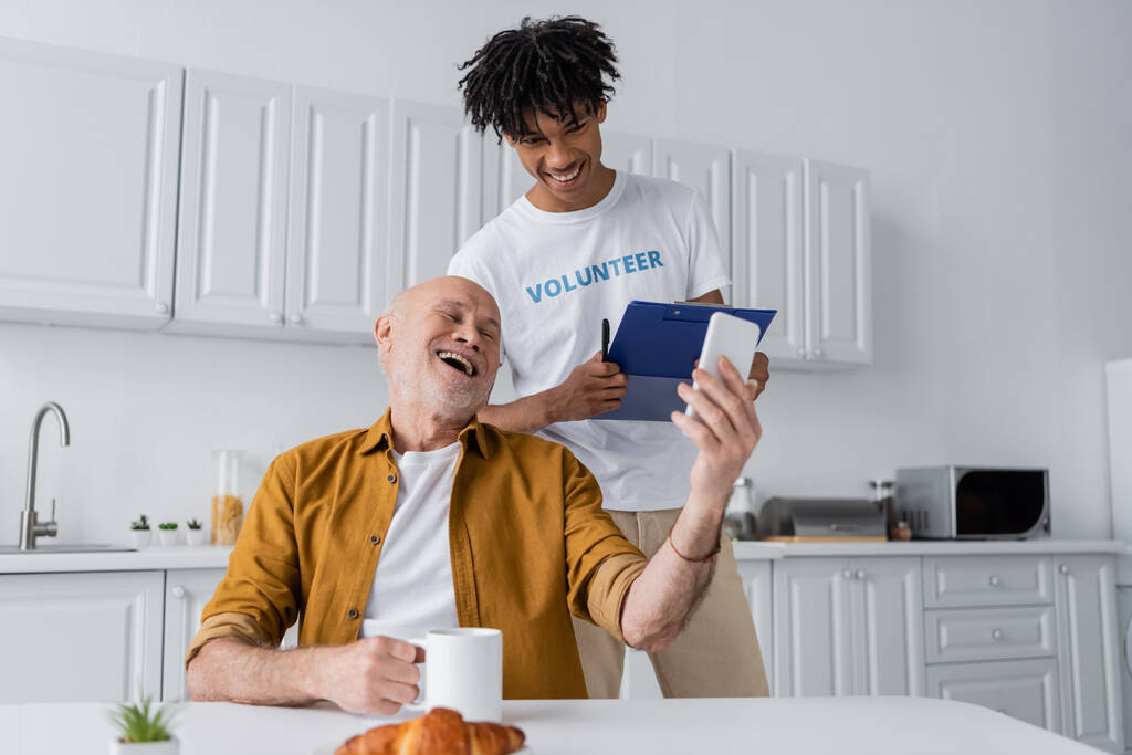 Positive senior man holding cellphone and cup near african american volunteer with clipboard in kitchen  - 写真・画像