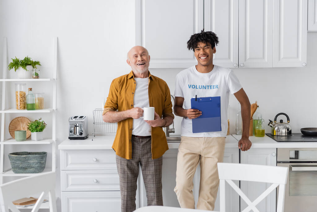 Cheerful african american volunteer with clipboard and elderly man looking at camera in kitchen  - Foto, imagen