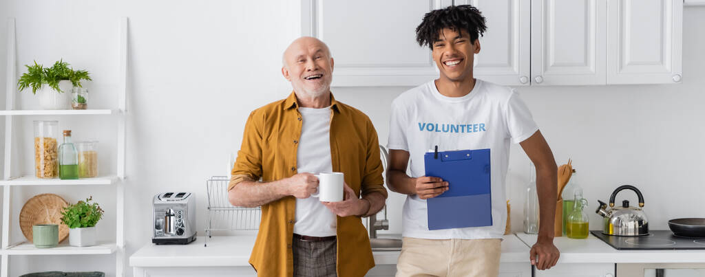 Happy senior man holding cup near african american volunteer with clipboard in kitchen, banner  - Foto, afbeelding