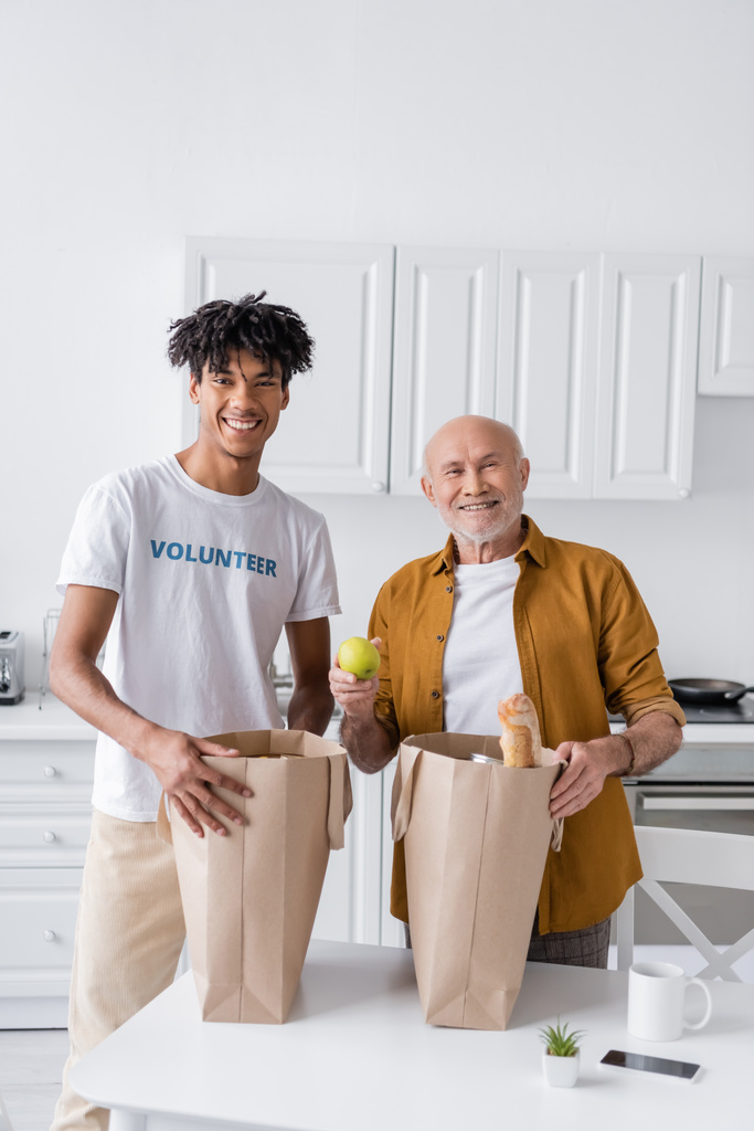 Позитивный африканский доброволец, стоящий рядом с пожилым мужчиной, и сумки с едой на кухне  - Фото, изображение