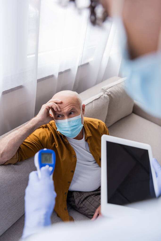 Старший чоловік в медичній масці сидить біля розмитої афроамериканської медсестри з цифровим планшетом і пірометром вдома
  - Фото, зображення