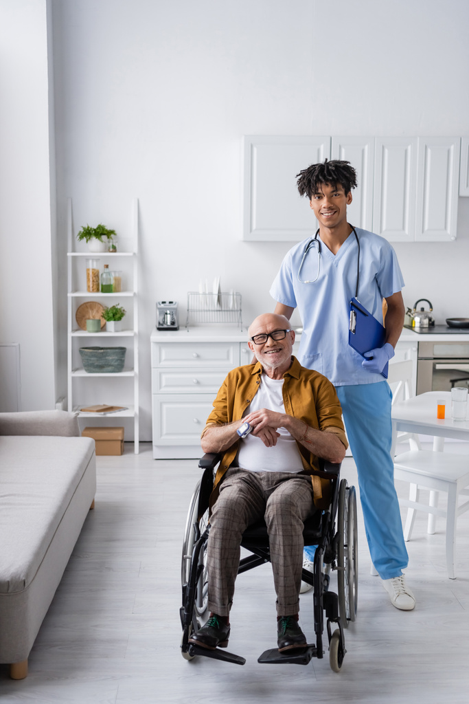 Smiling african american nurse with clipboard looking at camera near pensioner with pulse oximeter in wheelchair at home  - 写真・画像