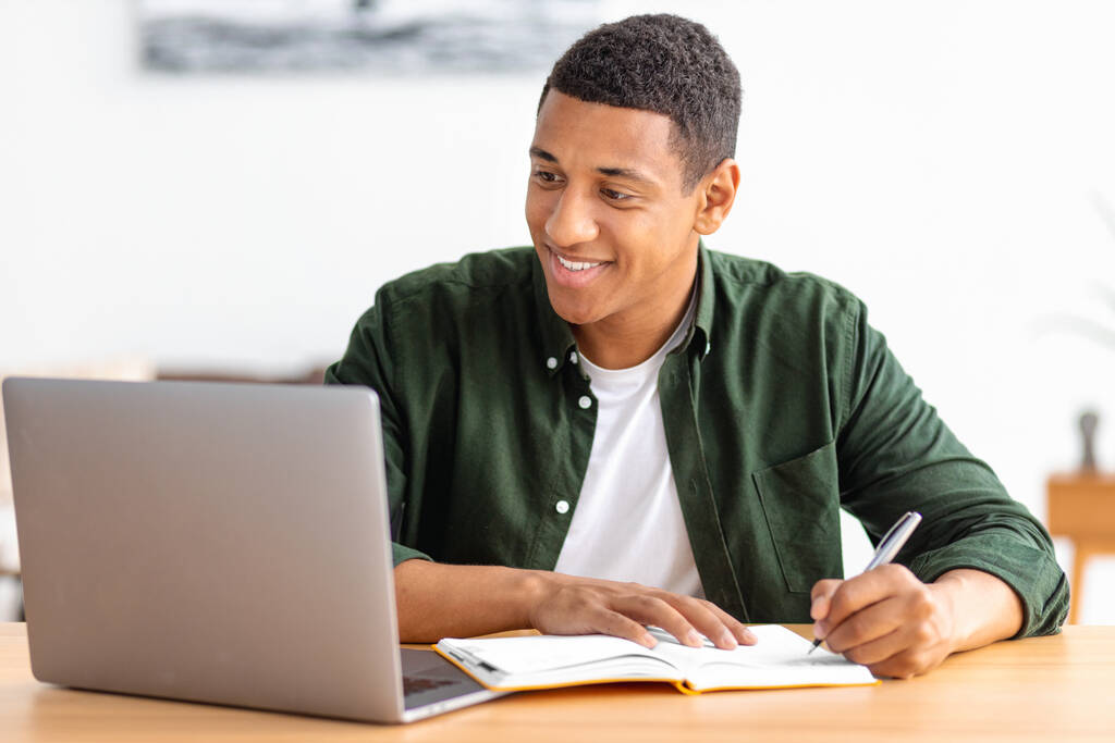 Smiling male student or freelancer studying remotely from home. African American male using laptop for online education, taking notes in a notebook. Online lesson, e-learning at home concept - Zdjęcie, obraz