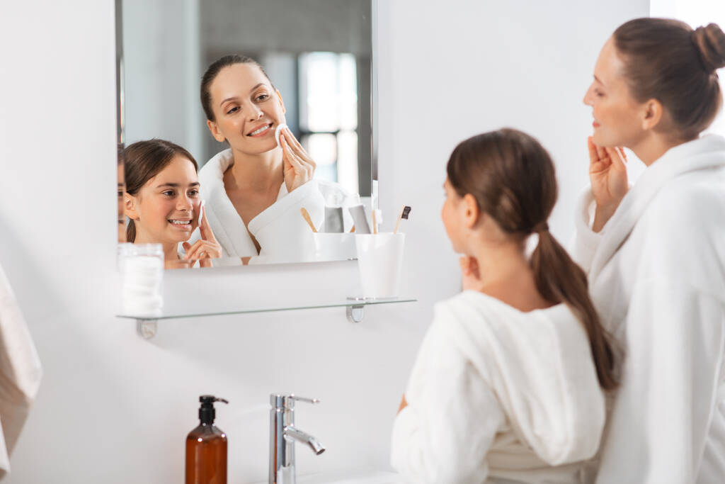 красота, гигиена, утренняя и народная концепция - счастливые улыбающиеся мать и дочь с хлопчатобумажными прокладками очищающие кожу лица и глядя на зеркало в ванной комнате - Фото, изображение