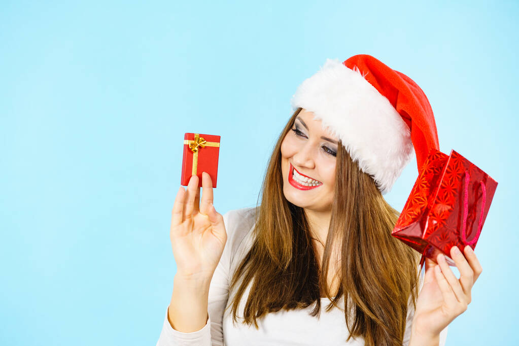 Christmas gifts. Happy woman wearing santa claus hat holding present red bag and small gift box with jewelery, on blue - Photo, Image