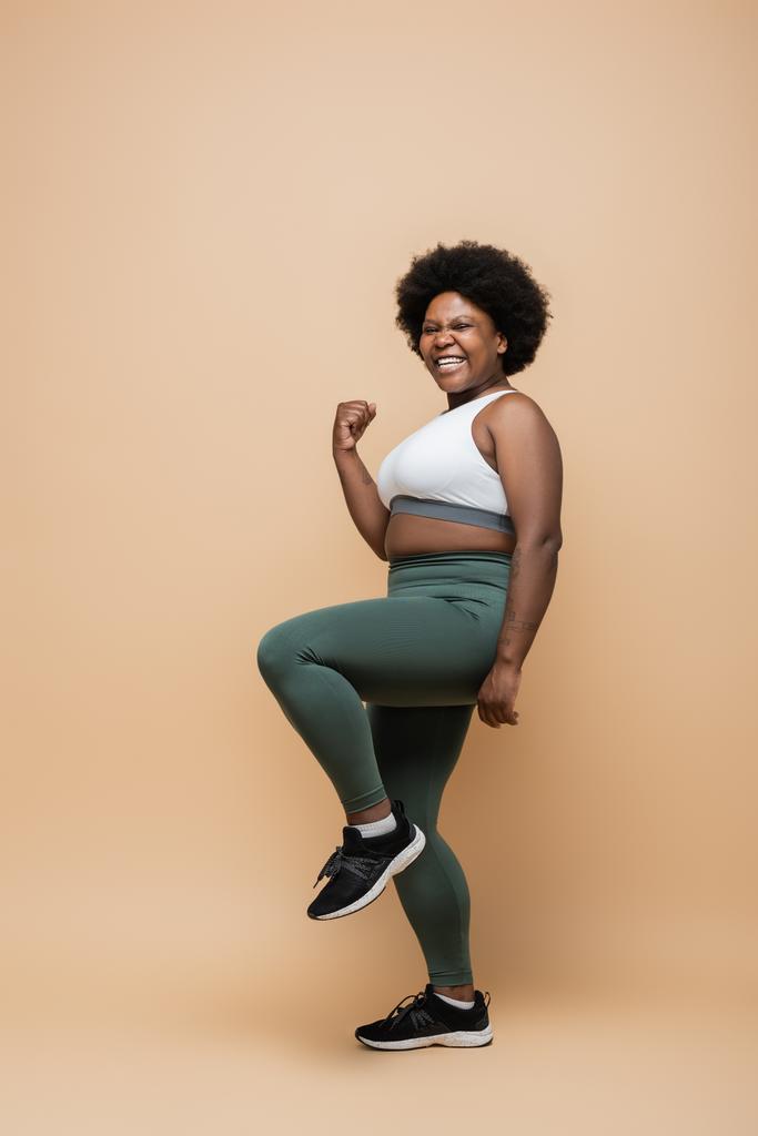 full length of happy african american plus size woman in sportswear showing yes gesture on beige - Foto, afbeelding