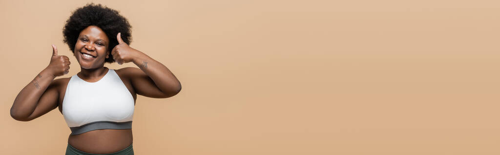щаслива афроамериканка плюс розмір жінка у верхній частині врожаю показує великі пальці вгору ізольовані на бежевому, банер
 - Фото, зображення
