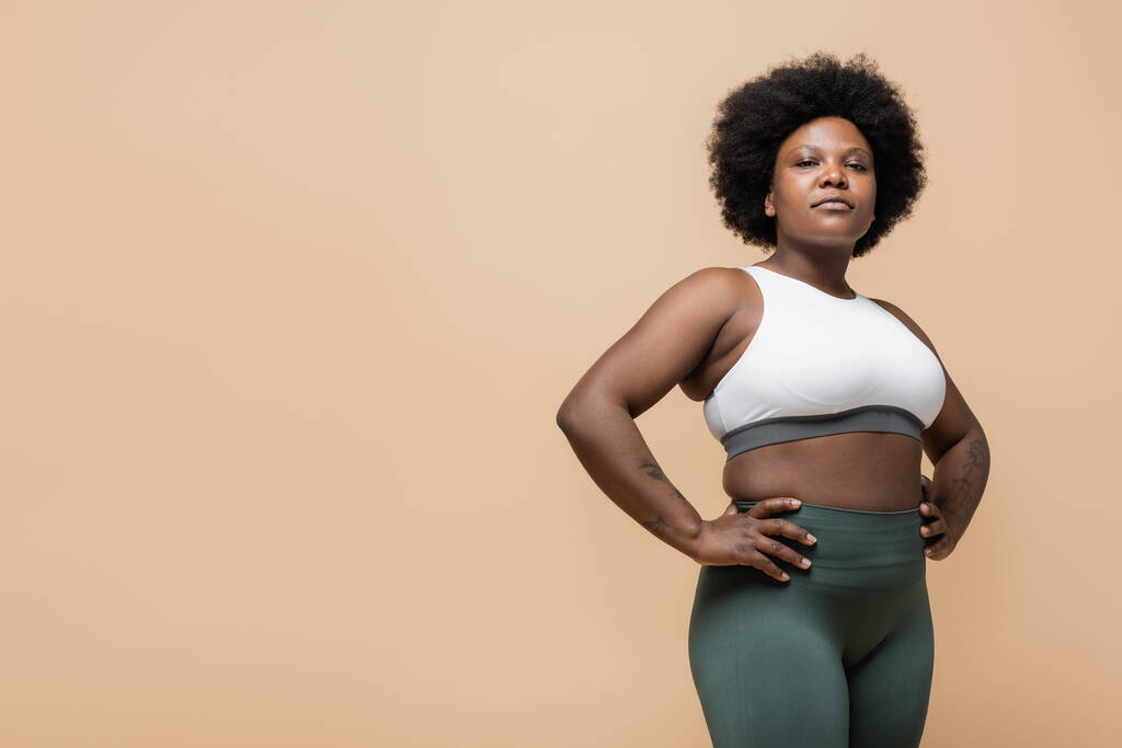 curly african american plus size woman in crop top standing with hands on hips isolated on beige - Foto, immagini