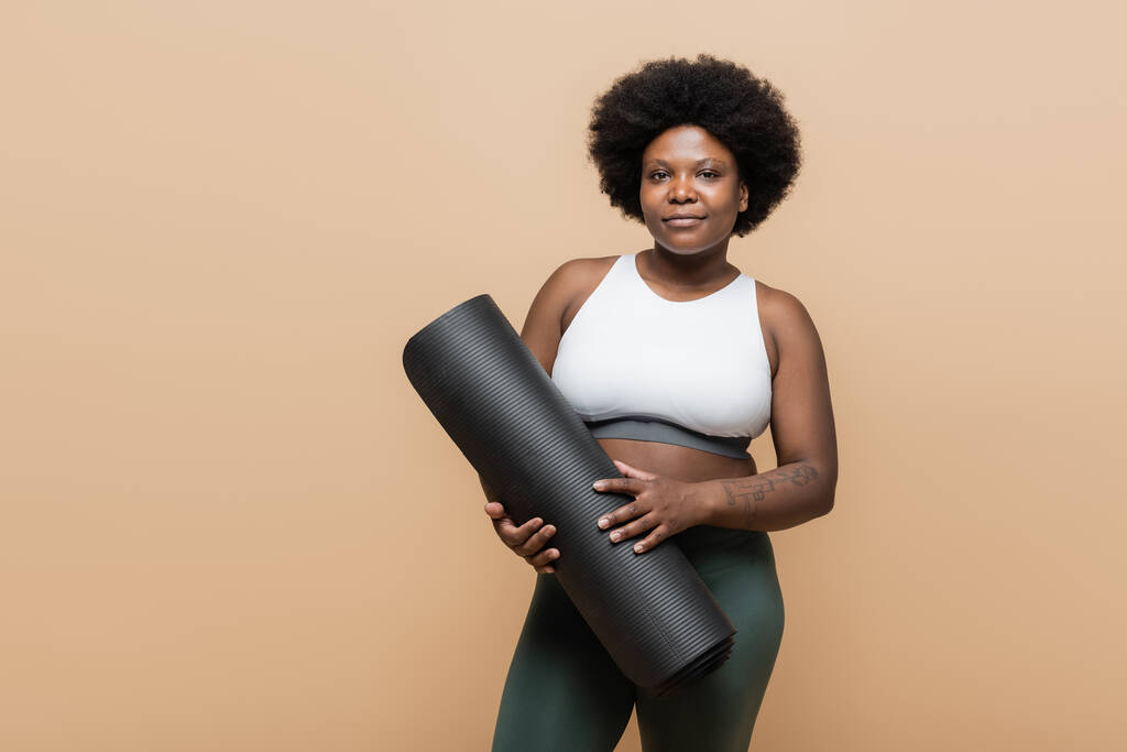 curly african american plus size woman in crop top holding fitness mat isolated on beige - Foto, Bild