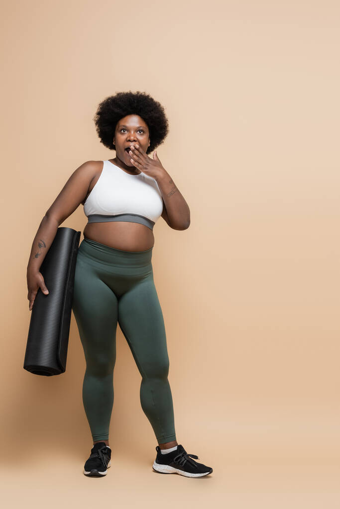 full length of shocked african american plus size woman holding fitness mat and covering opened mouth on beige - Foto, Bild