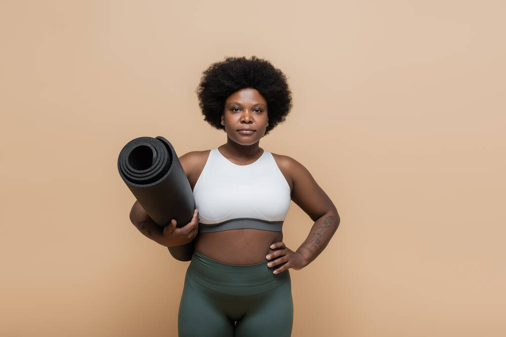 african american plus size woman in crop top holding fitness mat and standing with hand on hip isolated on beige - Foto, Bild