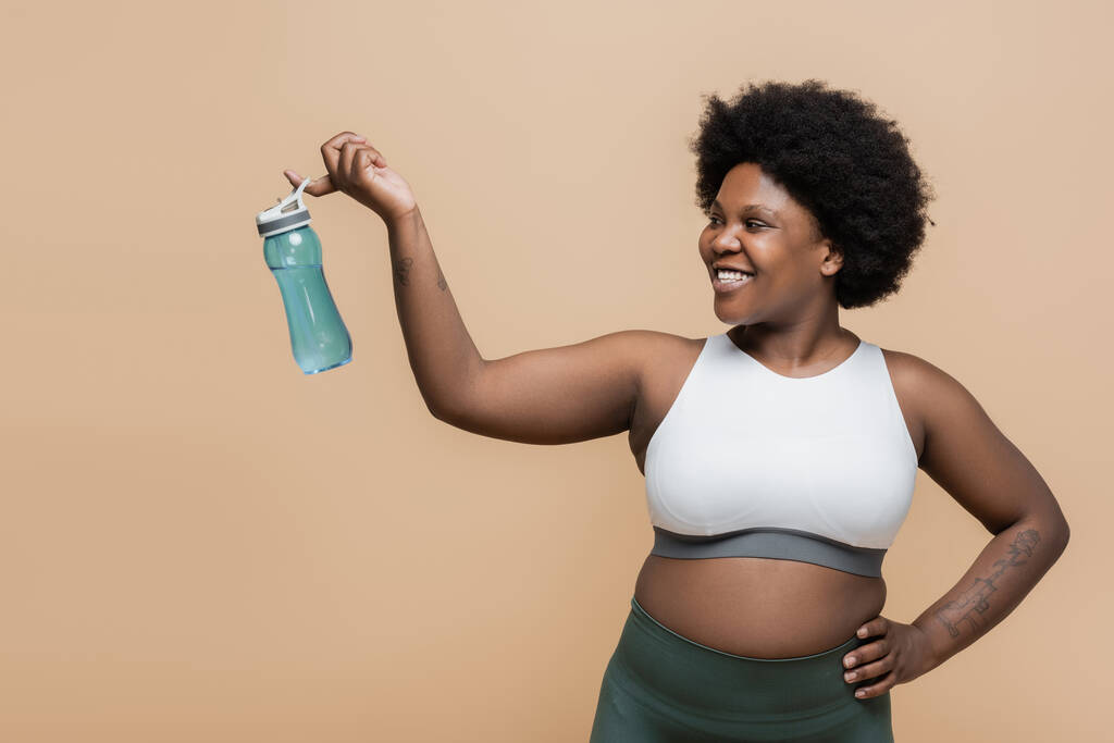 счастливый африканский американец плюс размер женщины в кукурузном топе держа спортивную бутылку на пальце изолированы на бежевый - Фото, изображение