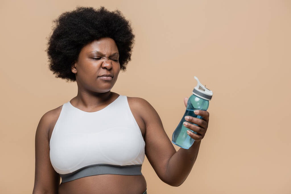 displeased african american plus size woman in crop top holding sports bottle with water isolated on beige - Foto, Bild