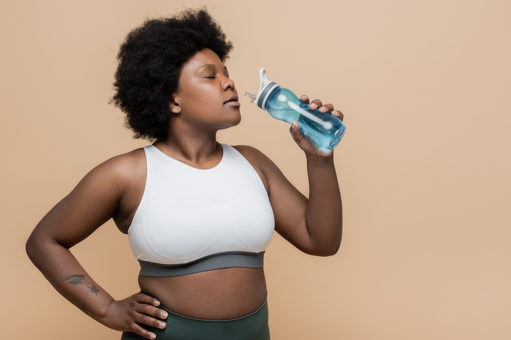Африканский американец плюс размер женщина держит спортивную бутылку с водой и стоя с рукой на бедре изолированы на бежевый - Фото, изображение