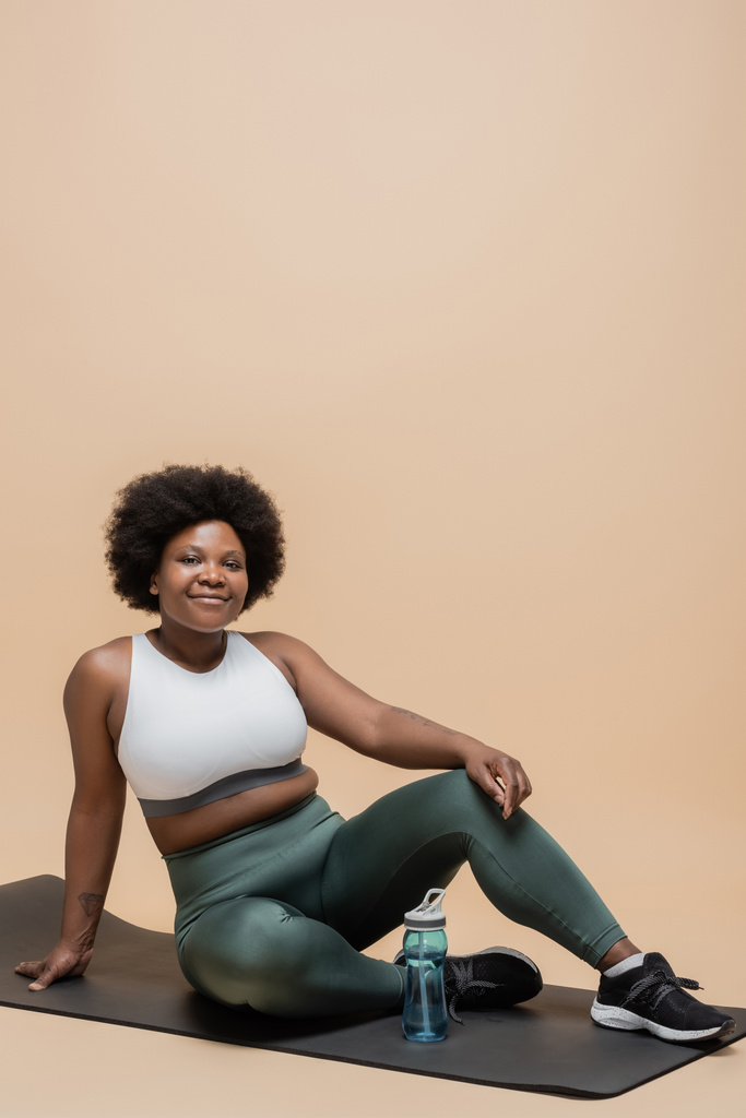 positive african american plus size woman in sportswear sitting on fitness mat near sports bottle on beige - Foto, Bild