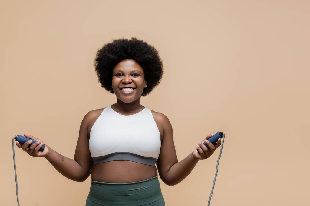 smiling african american plus size woman in sportswear holding skipping rope isolated on beige  - Foto, imagen