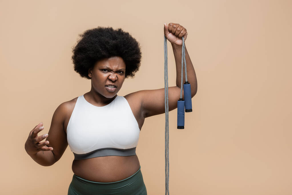 недовольный африканский американец плюс размер женщины в спортивной одежде держа скакалку изолированы на бежевый  - Фото, изображение