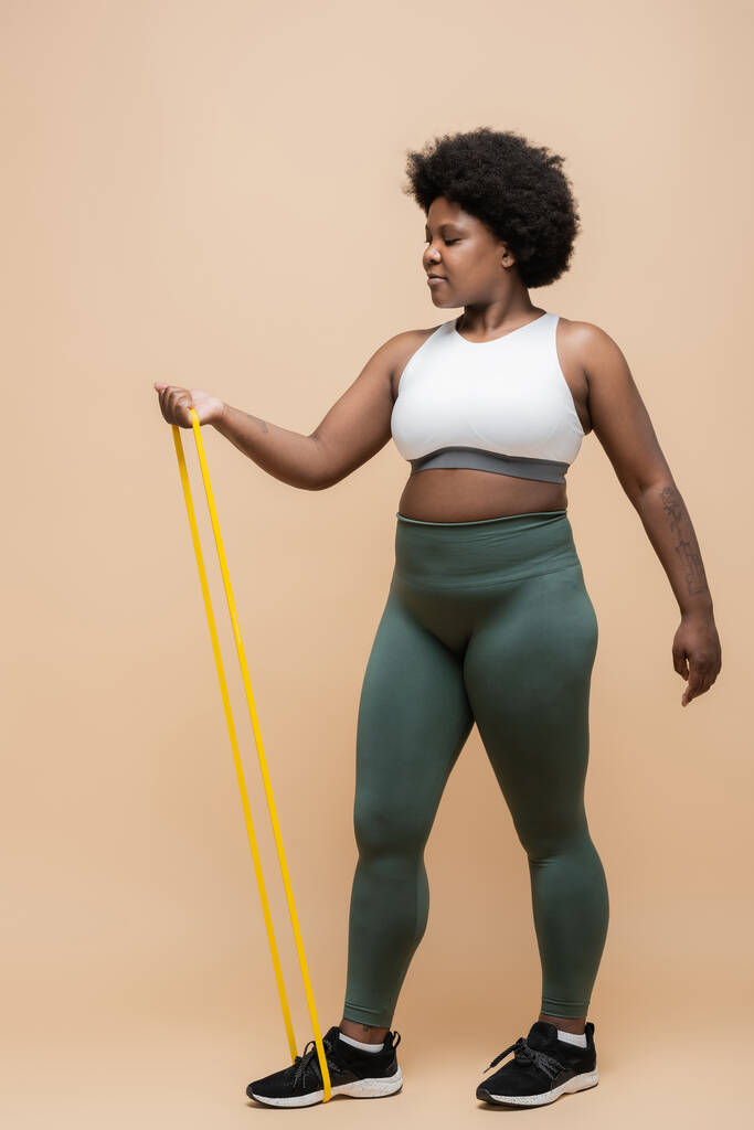 full length of curly african american plus size woman in sportwear exercising with resistance band on beige - Foto, afbeelding