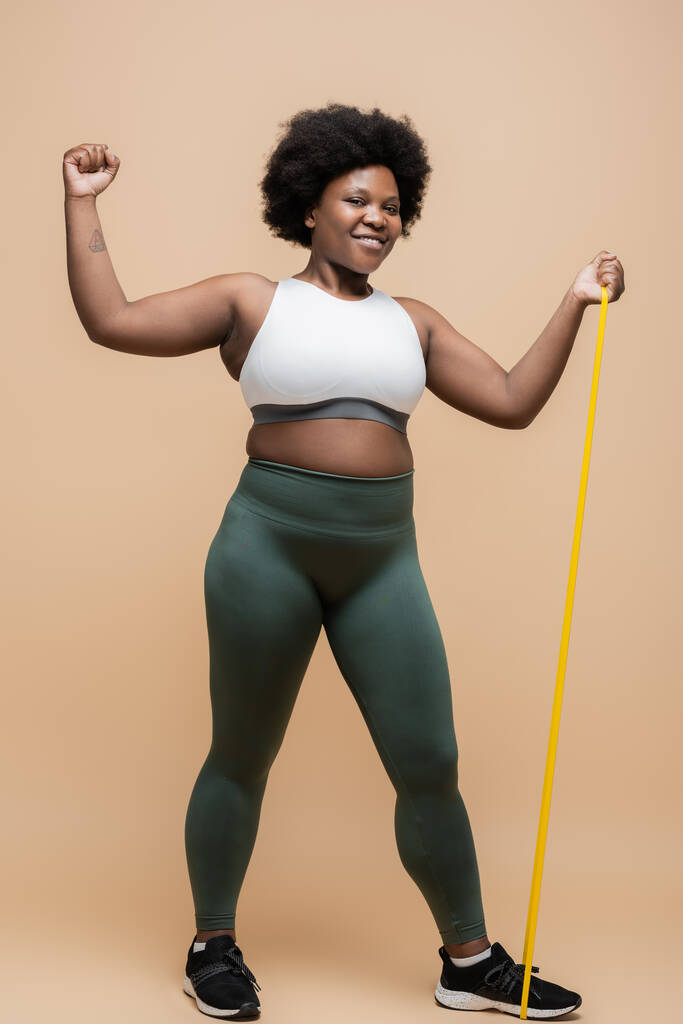 full length of joyful african american plus size woman in sportwear exercising with resistance band on beige - Valokuva, kuva
