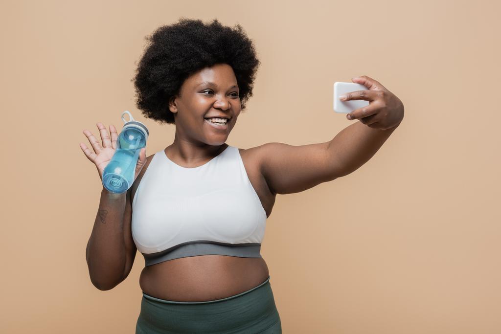 щаслива афроамериканка плюс розмір жінка тримає спортивну пляшку і махає рукою під час відеодзвінка на смартфон ізольовано на бежевому
 - Фото, зображення