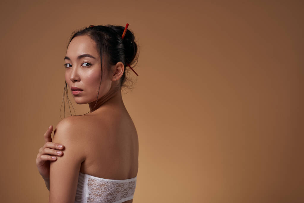 Side view of serious elegant asian girl hugging herself and looking at camera. Beautiful young brunette slim woman wearing tank top. Female beauty. Isolated on orange background in studio. Copy space - Zdjęcie, obraz