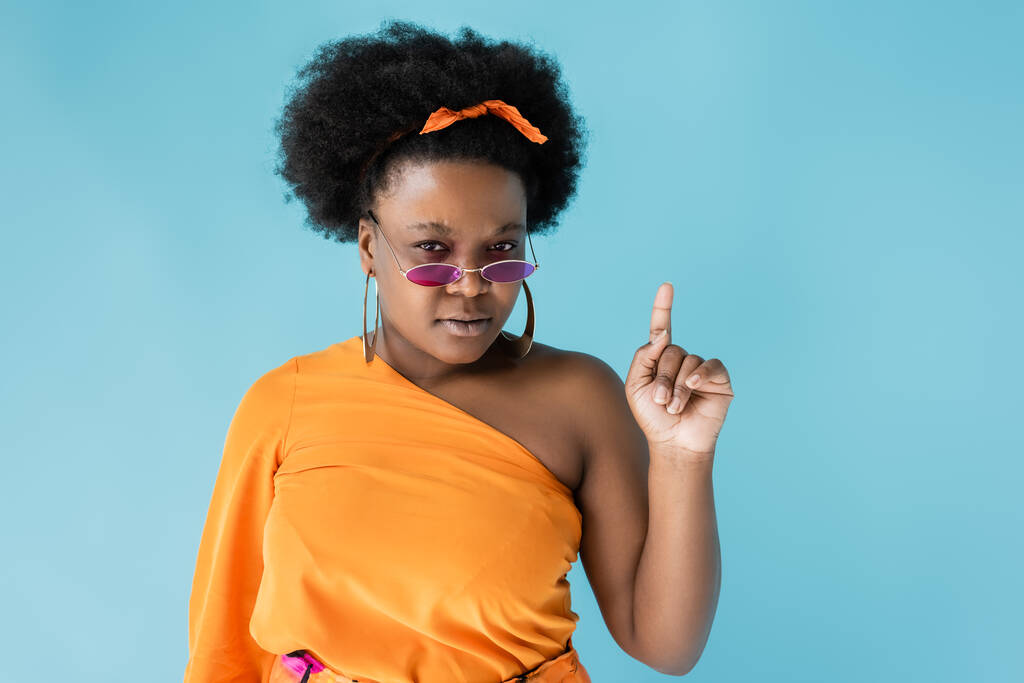 curly african american plus size woman in sunglasses and hoop earrings pointing up isolated on blue - Foto, afbeelding