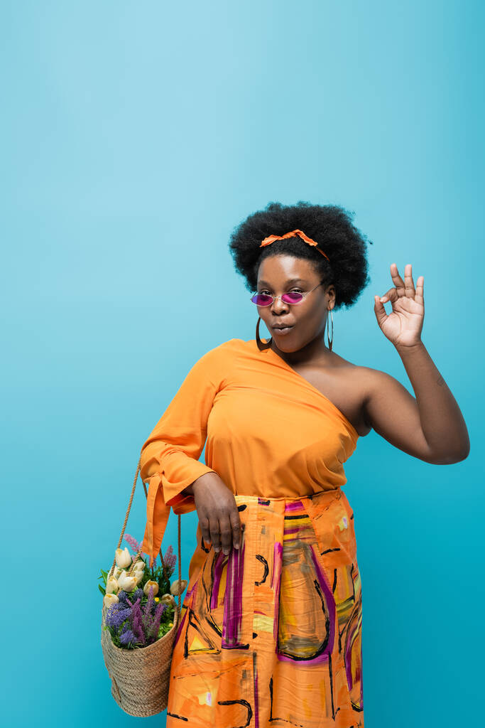 curly african american body positive woman in sunglasses holding straw bag with flowers and showing ok isolated on blue - Fotó, kép