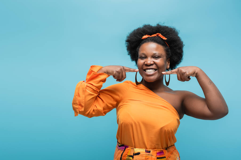 cheerful african american body positive woman in orange dress and hoop earrings pointing at cheeks isolated on blue - Foto, Bild
