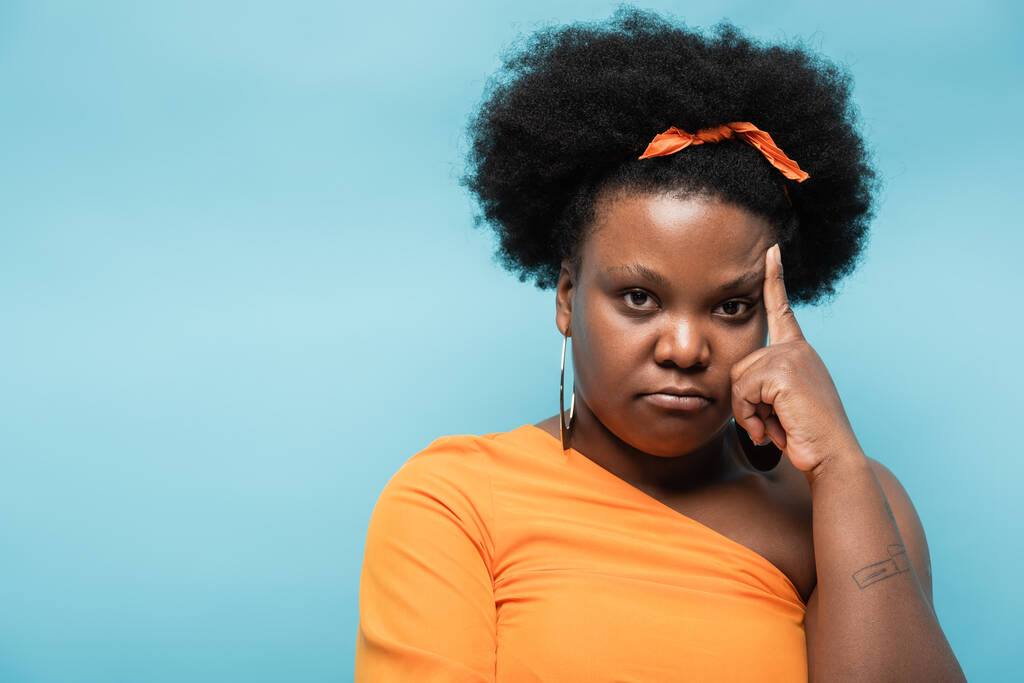 displeased african american body positive woman in orange dress and hoop earrings isolated on blue - 写真・画像