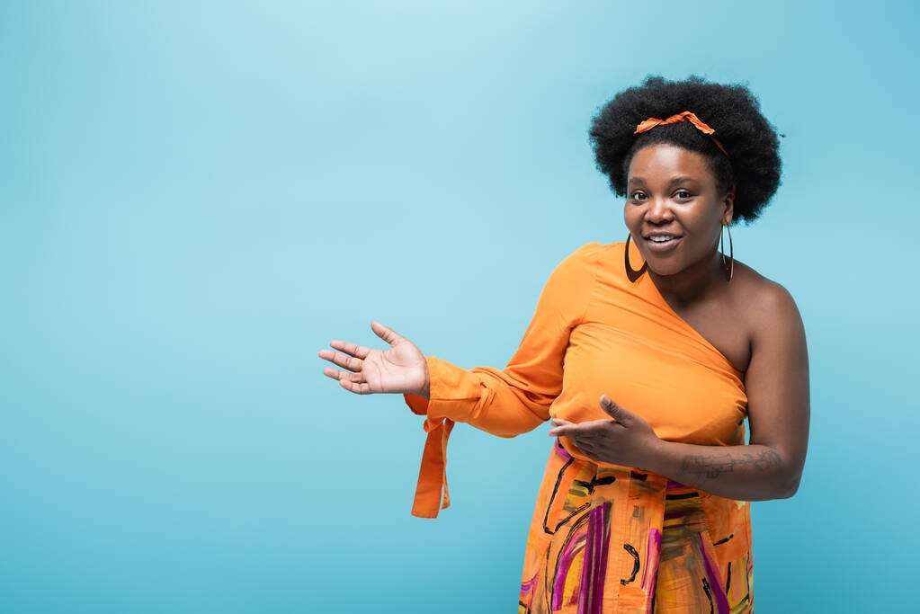 smiling african american body positive woman in orange dress and hoop earrings pointing with hands isolated on blue - Foto, Imagen