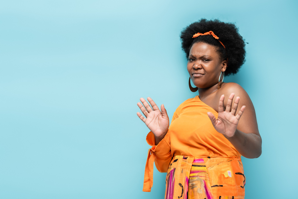displeased african american body positive woman in hoop earrings and orange dress showing no gesture isolated on blue - 写真・画像