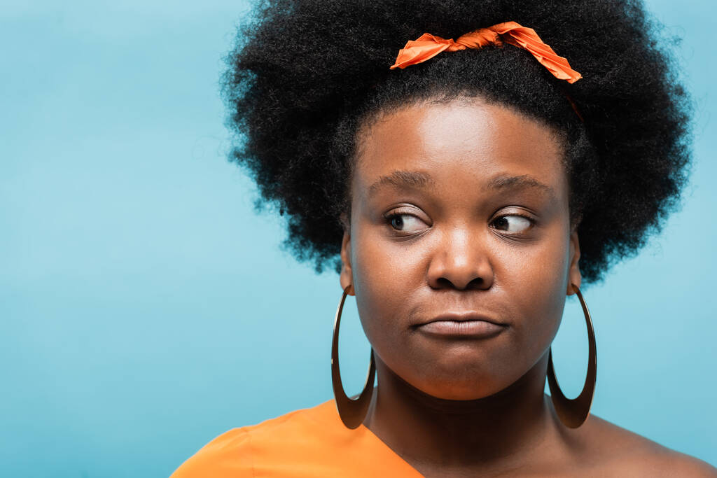 portrait of curly african american body positive woman in hoop earrings looking away isolated on blue - Zdjęcie, obraz