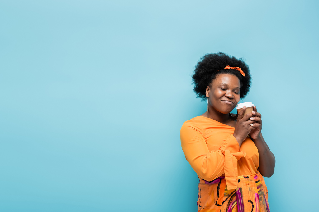 pleased african american body positive woman in hoop earrings holding paper cup isolated on blue - Foto, Bild