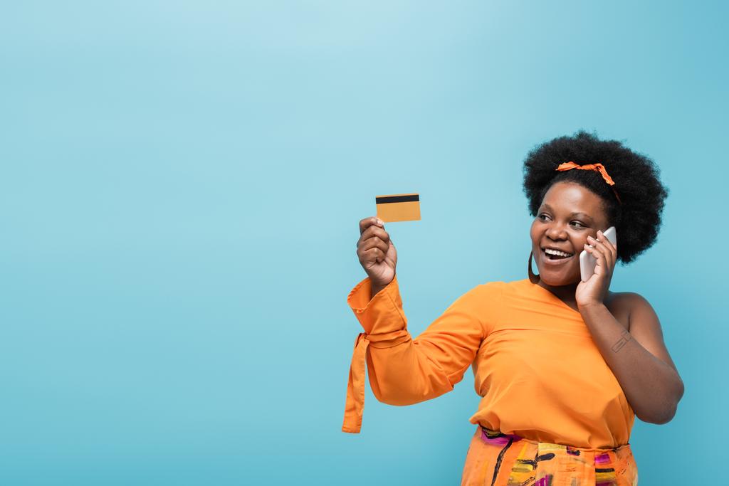 happy african american body positive woman in hoop earrings holding credit card and talking on smartphone isolated on blue - Foto, Imagem