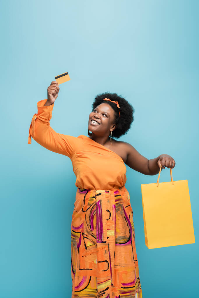 cheerful african american body positive woman in orange dress holding shopping bag and credit card isolated on blue - Foto, afbeelding