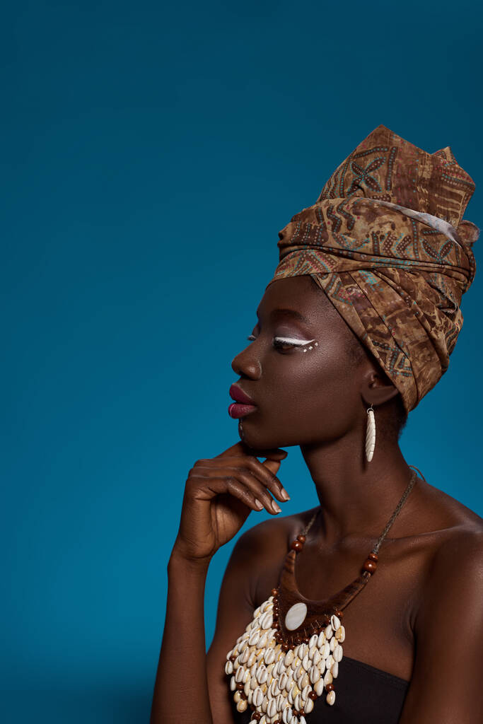 Profile off fashionable black girl thinking about something. Pretty young woman wearing traditional african outfit and accessories. Female beauty. Isolated on blue background. Studio shoot. Copy space - Zdjęcie, obraz