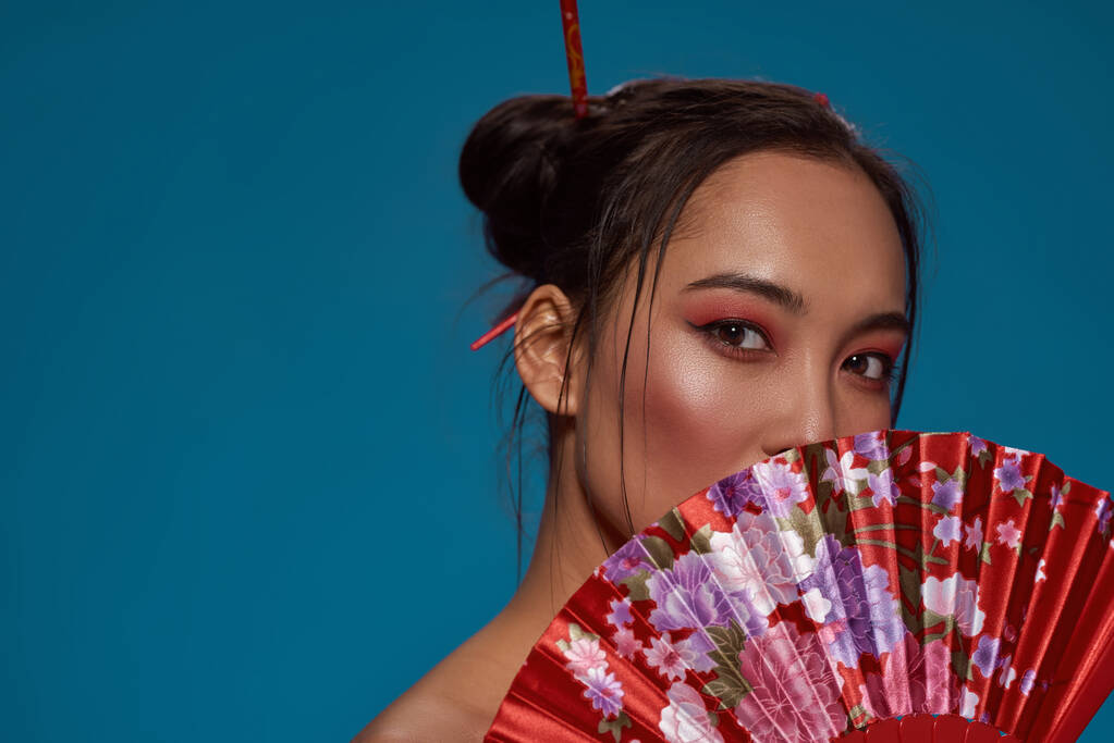 Partial of elegant asian girl covering part of face with traditional handheld fan. Beautiful young brunette woman look at camera. Female beauty. Isolated on blue background. Studio shoot. Copy space - Valokuva, kuva