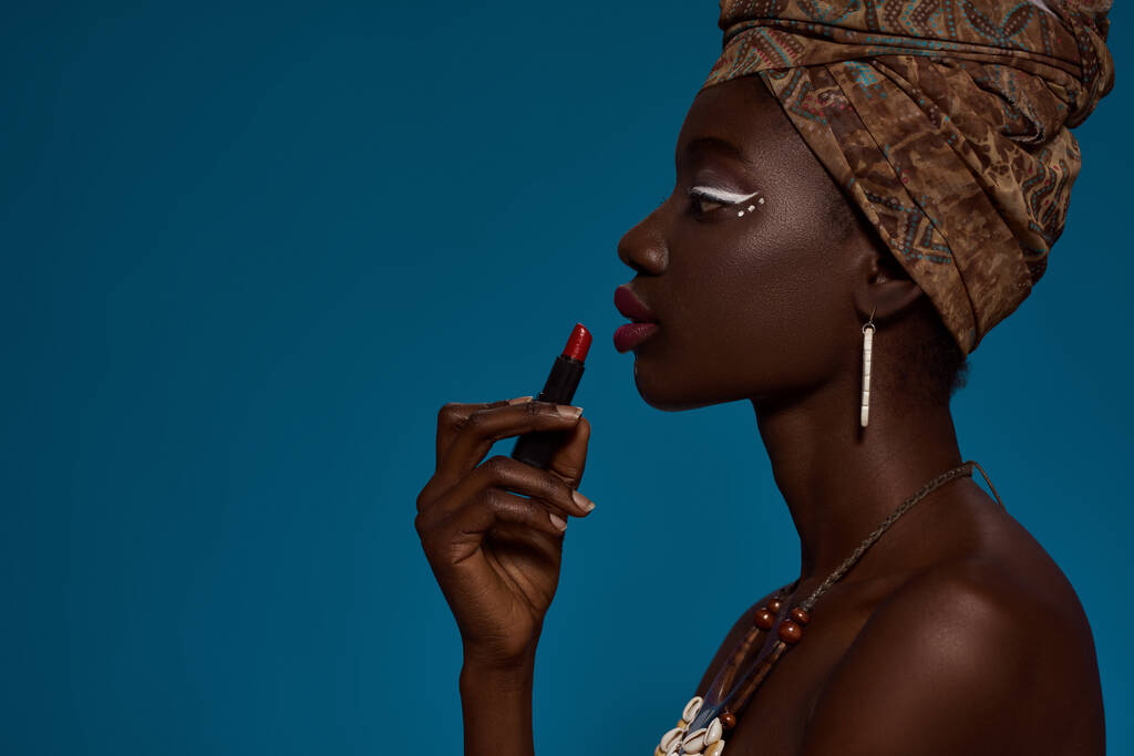 Partial side view of stylish black girl painting lips with lipstick. Attractive young woman wearing traditional african outfit and accessories. Female beauty. Blue background. Studio shoot. Copy space - Foto, Imagem