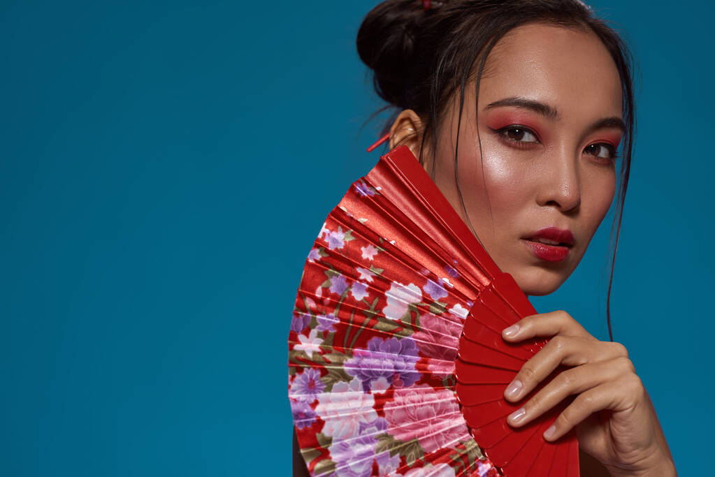 Cropped of serious fashionable asian girl posing with traditional handheld fan and looking at camera. Pretty young brunette woman. Female beauty. Isolated on blue background. Studio shoot. Copy space - Fotografie, Obrázek