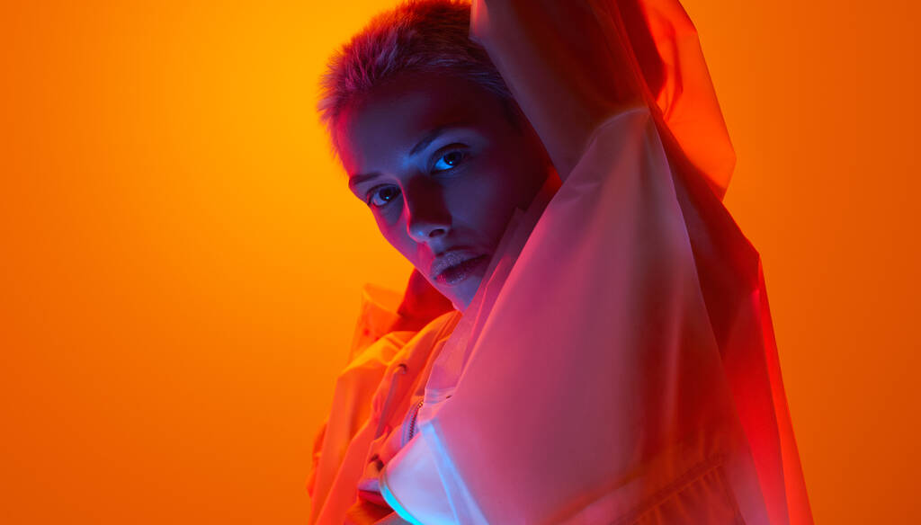 Side view of confident young female model with short blond hair in raincoat, standing against orange background with raised arms and looking at camera in neon light - Valokuva, kuva