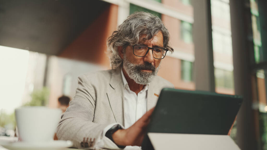 Mature businessman with beard in eyeglasses wearing gray jacket, working on tablet, sitting in an outdoor cafe. Successful man sitting at table in cafe in nature Mobile office freelancer - Fotoğraf, Görsel