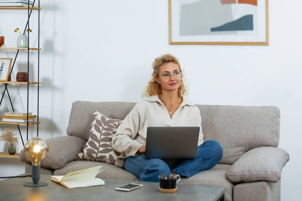 Portrait of adult blonde woman wearing a sweater and glasses sitting on the couch at home and working on a laptop. Background, copy space, close up. - Fotoğraf, Görsel