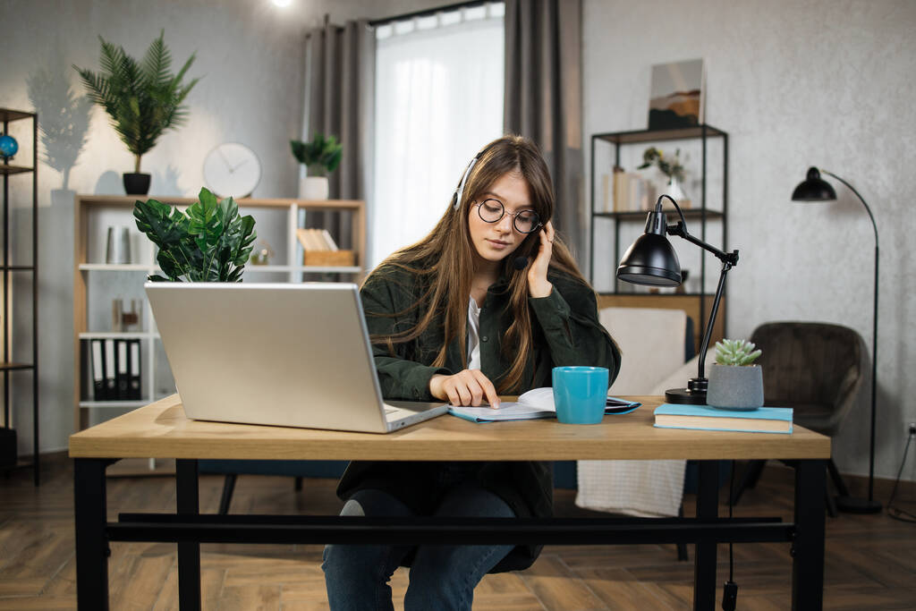 Νεαρή καυκάσια γυναίκα με ακουστικά που κάθεται στο γραφείο με φορητό υπολογιστή και μιλάει κατά τη διάρκεια βιντεοκλήσης. Επιχειρηματίας που χρησιμοποιεί σύγχρονες συσκευές για online συνομιλία στο χώρο εργασίας. - Φωτογραφία, εικόνα