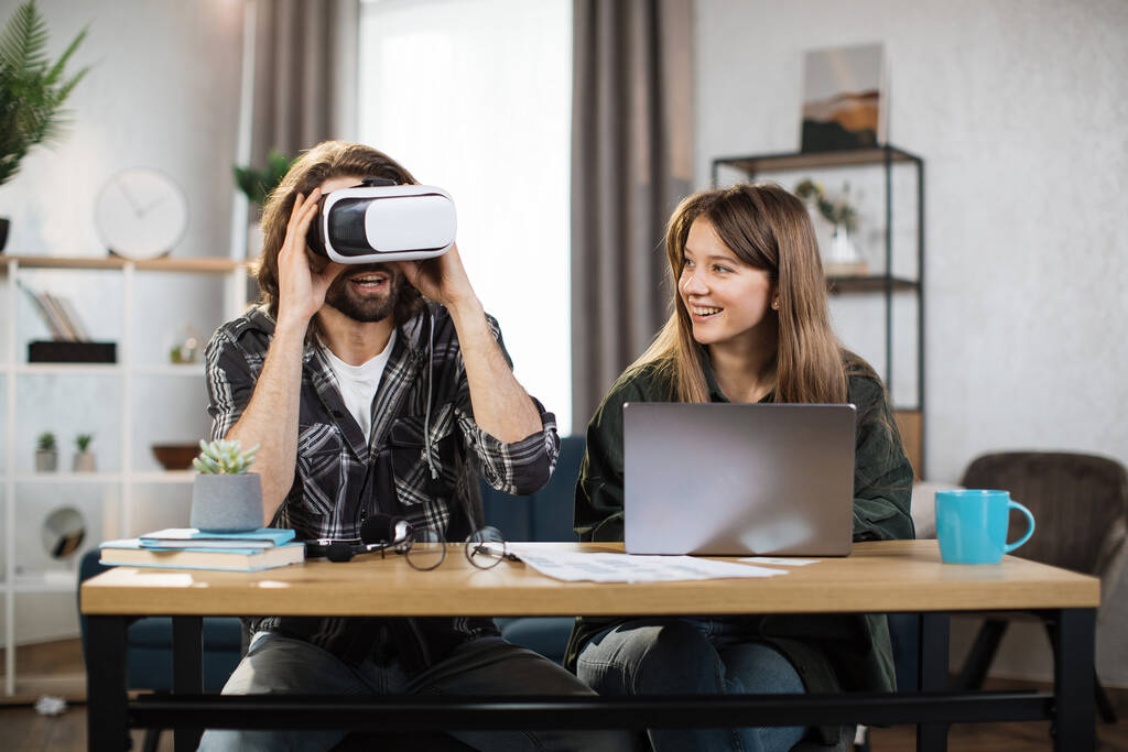 Innoissaan nuoret yllättynyt ilme, pelejä kotona ja käyttämällä VR lasit ja kannettava tietokone. Onnellinen mies käyttää VR-kuulokkeita, kun taas kaunis tyttö käyttää kannettavaa tietokonetta ja hymyilee - Valokuva, kuva